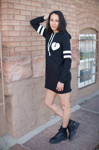 Woman wearing a black hoodie dress with white heart design against a brick wall.